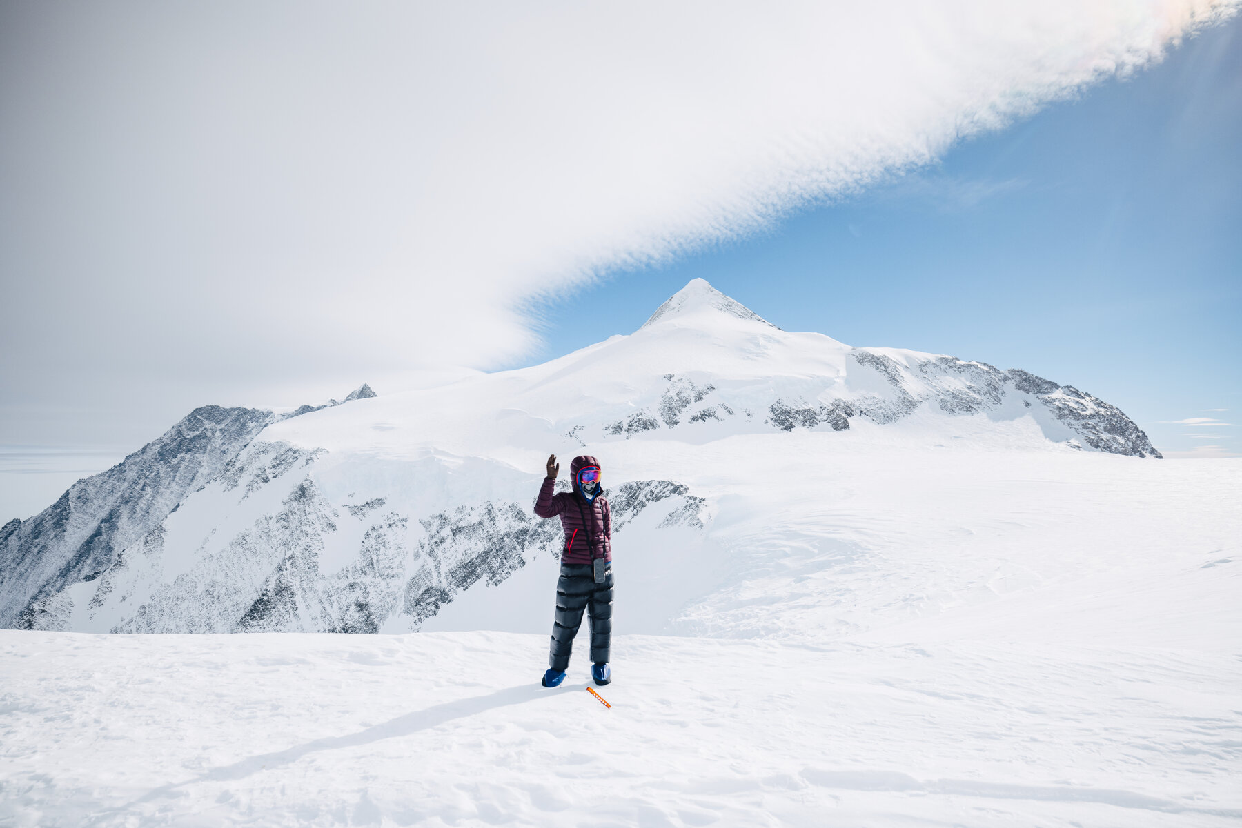 Solo climber in a maroon down suit on the High Camp plateau giving a wave, with a perfect snow pyramid summit rising behind under a wisp of cloud