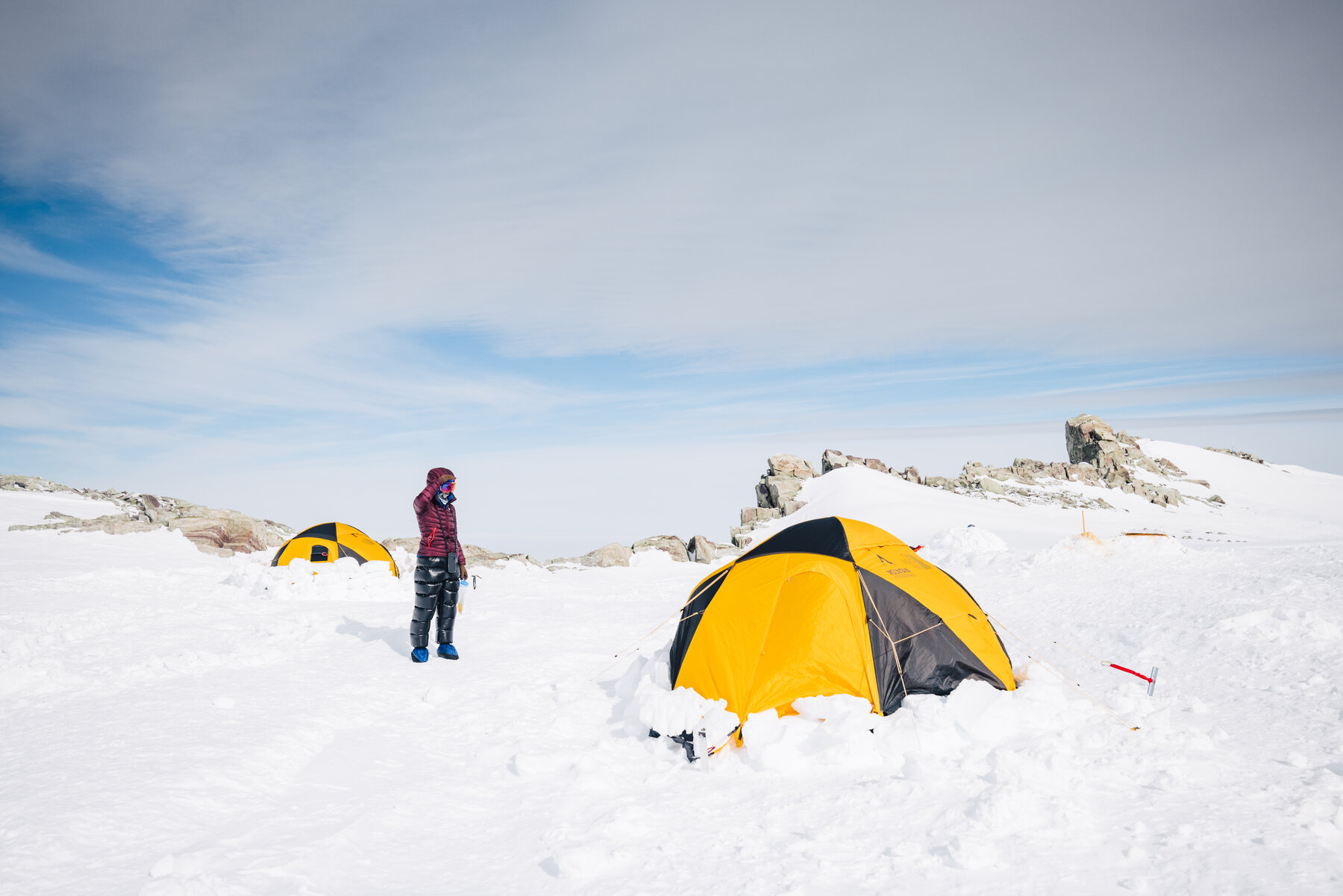 Climber in a maroon puffy beside a yellow expedition tent on the rocky High Camp plateau, scattered boulders behind