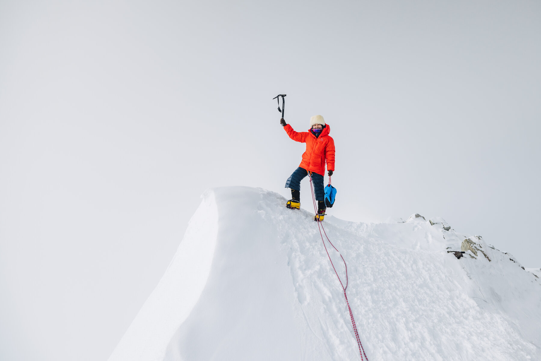 Lone climber in an orange down jacket standing on the corniced summit ridge with ice axe held high in triumph
