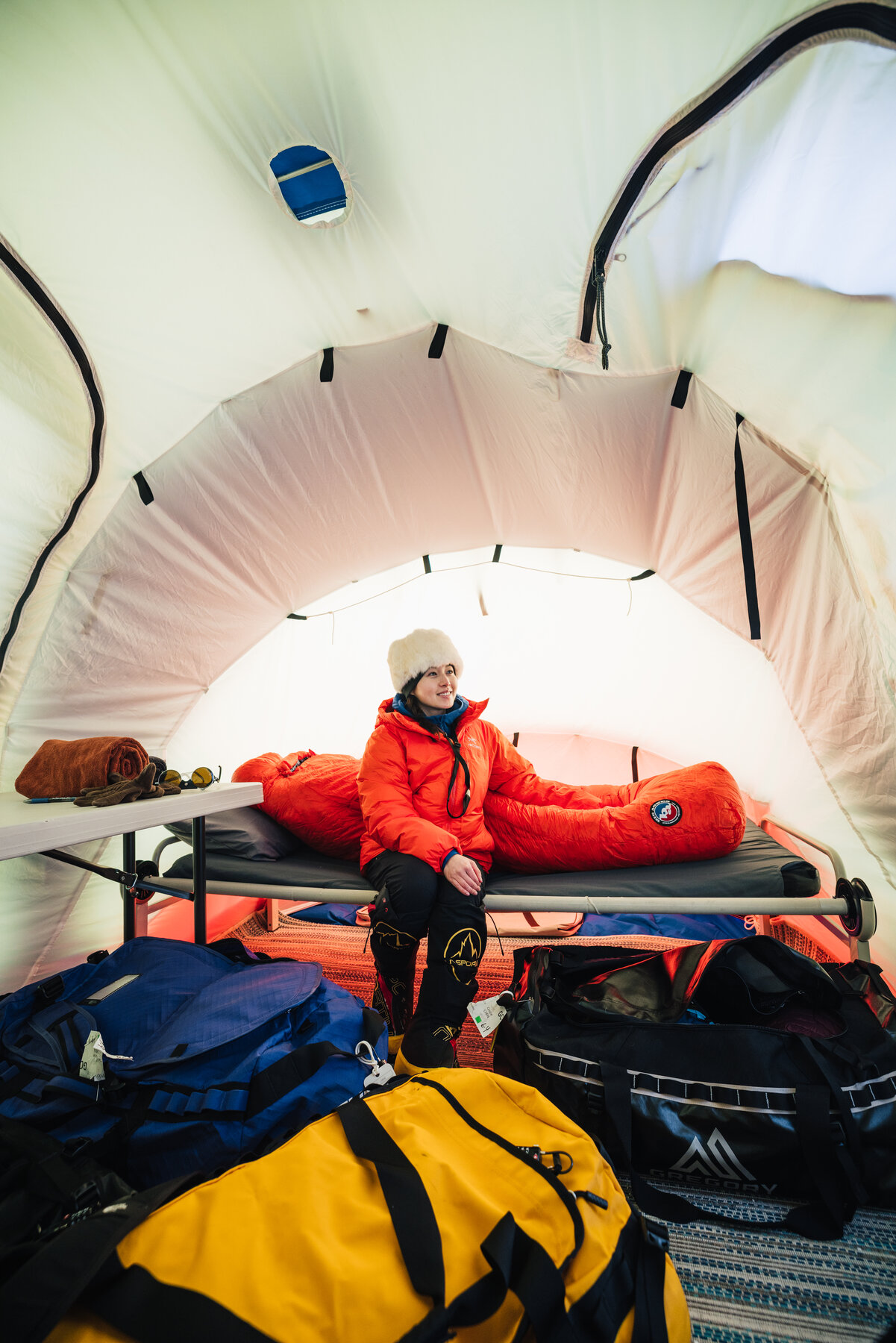 Climber in a red jacket sitting on a cot inside a Union Glacier sleeping tent with duffels and gear piled at the foot of the bed