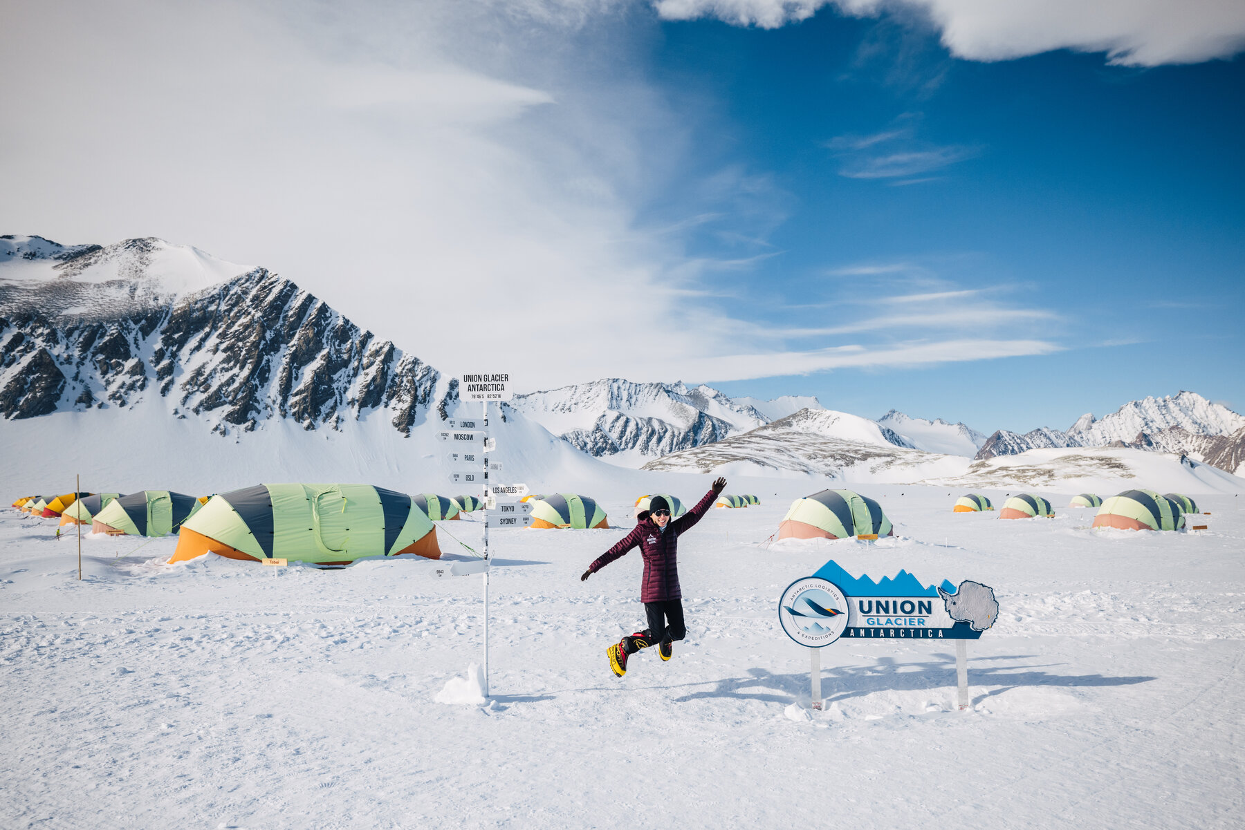 Climber leaping in the air beside the Union Glacier Camp welcome sign with dome tents and snowy peaks behind