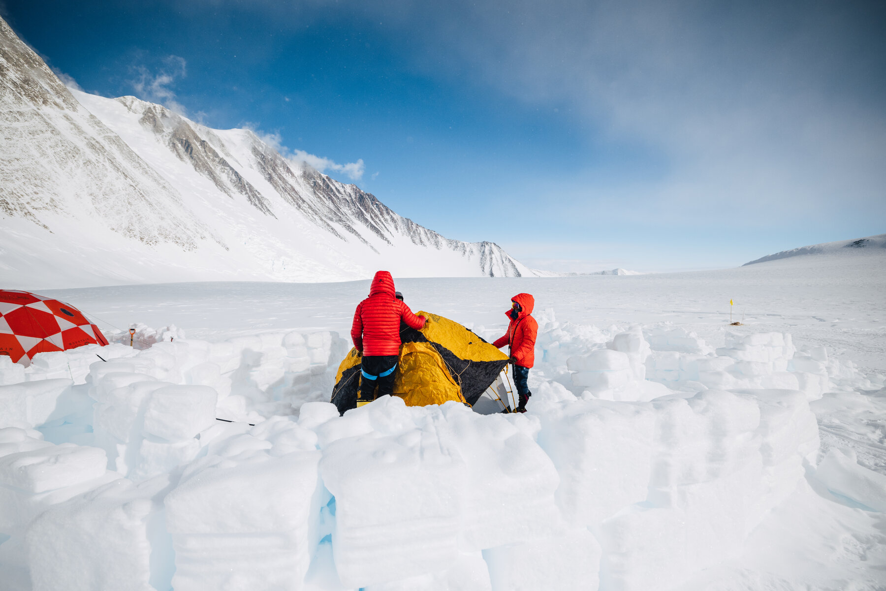 Two climbers in red down jackets pitching a yellow dome tent inside a fortress of snow-block walls at Low Camp, Vinson's west face rising behind