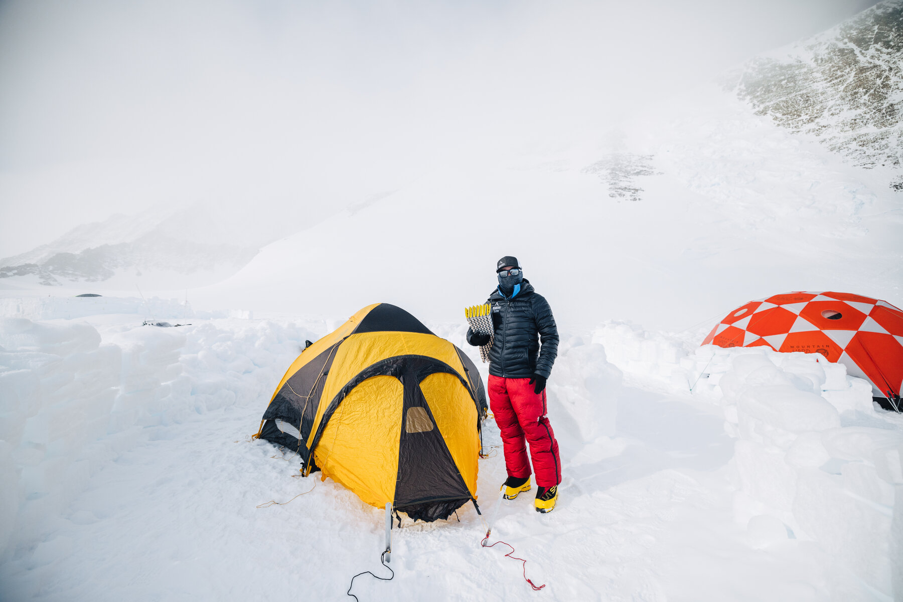 Climber in a black down jacket and red pants beside a yellow dome tent at Low Camp — snow-block walls in the foreground, a red checkerboard Mountain Hardwear tent on the right