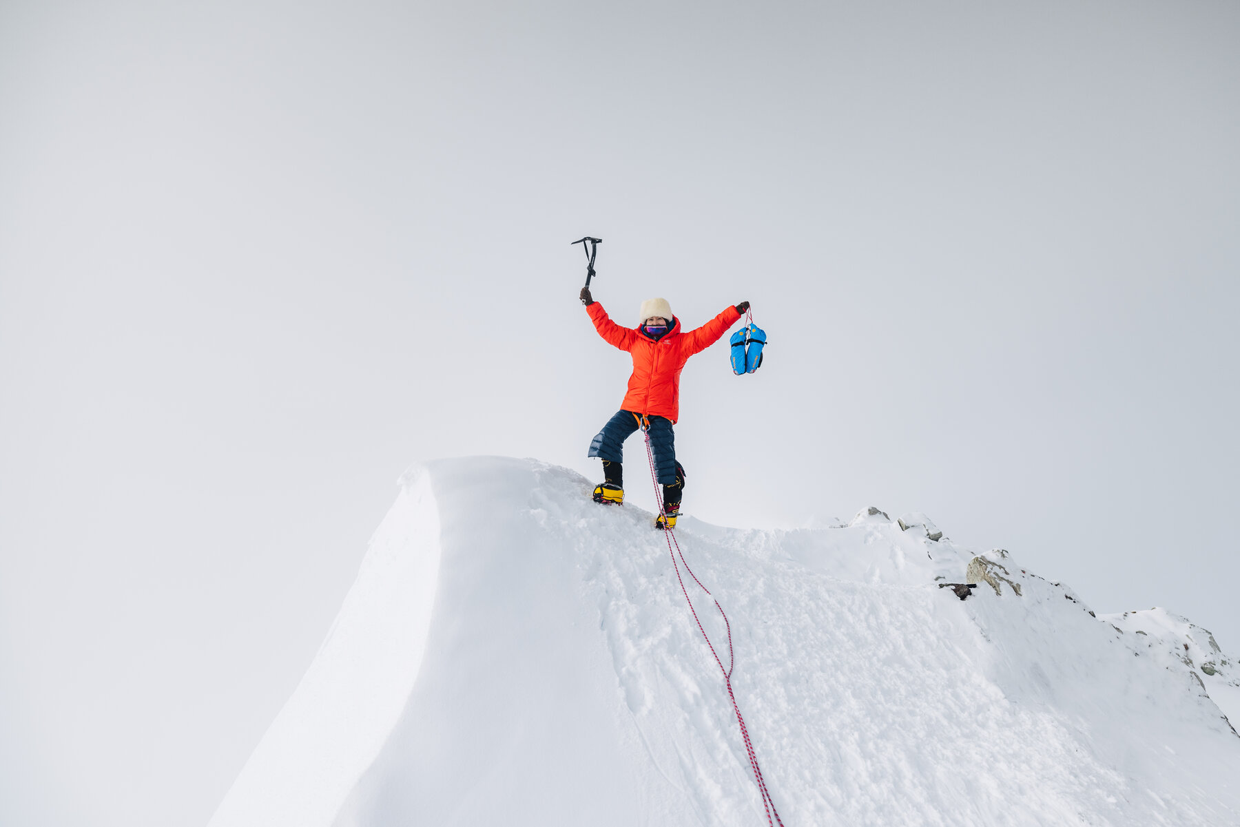 Climber in an orange down jacket standing on the corniced summit of Vinson with both arms and ice axe raised