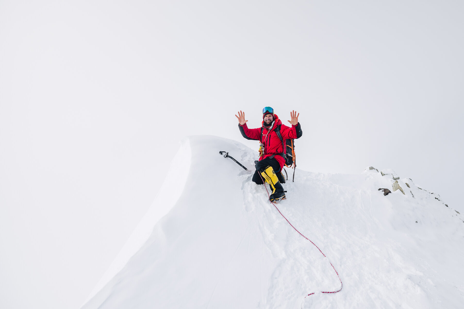 Climber in a red jacket and yellow boots standing on the corniced summit with both hands raised
