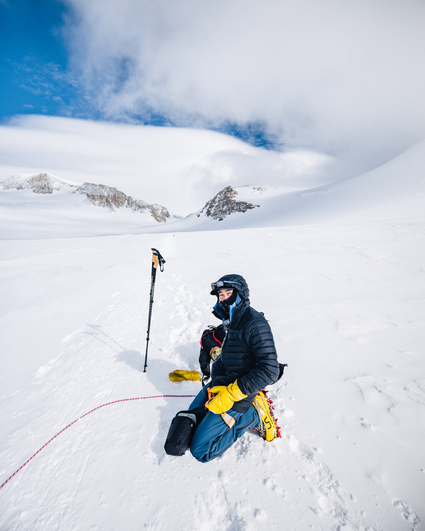 Climber in a black down jacket kneeling in the snow on summit day with trekking poles planted alongside, blue sky and peaks behind