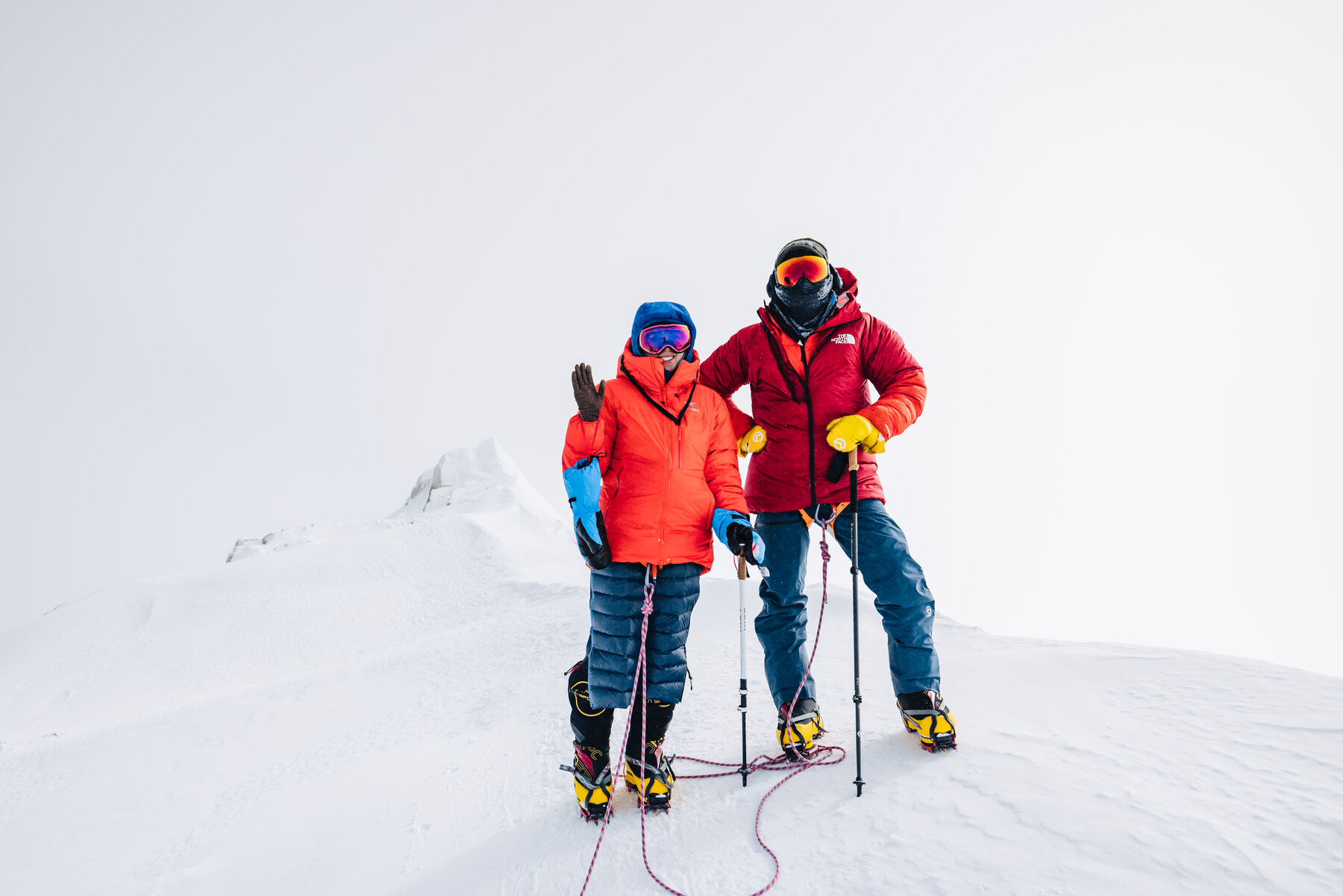 Two climbers in orange and red down jackets standing together on the Vinson summit, ice axe and poles in hand, white-out cloud behind