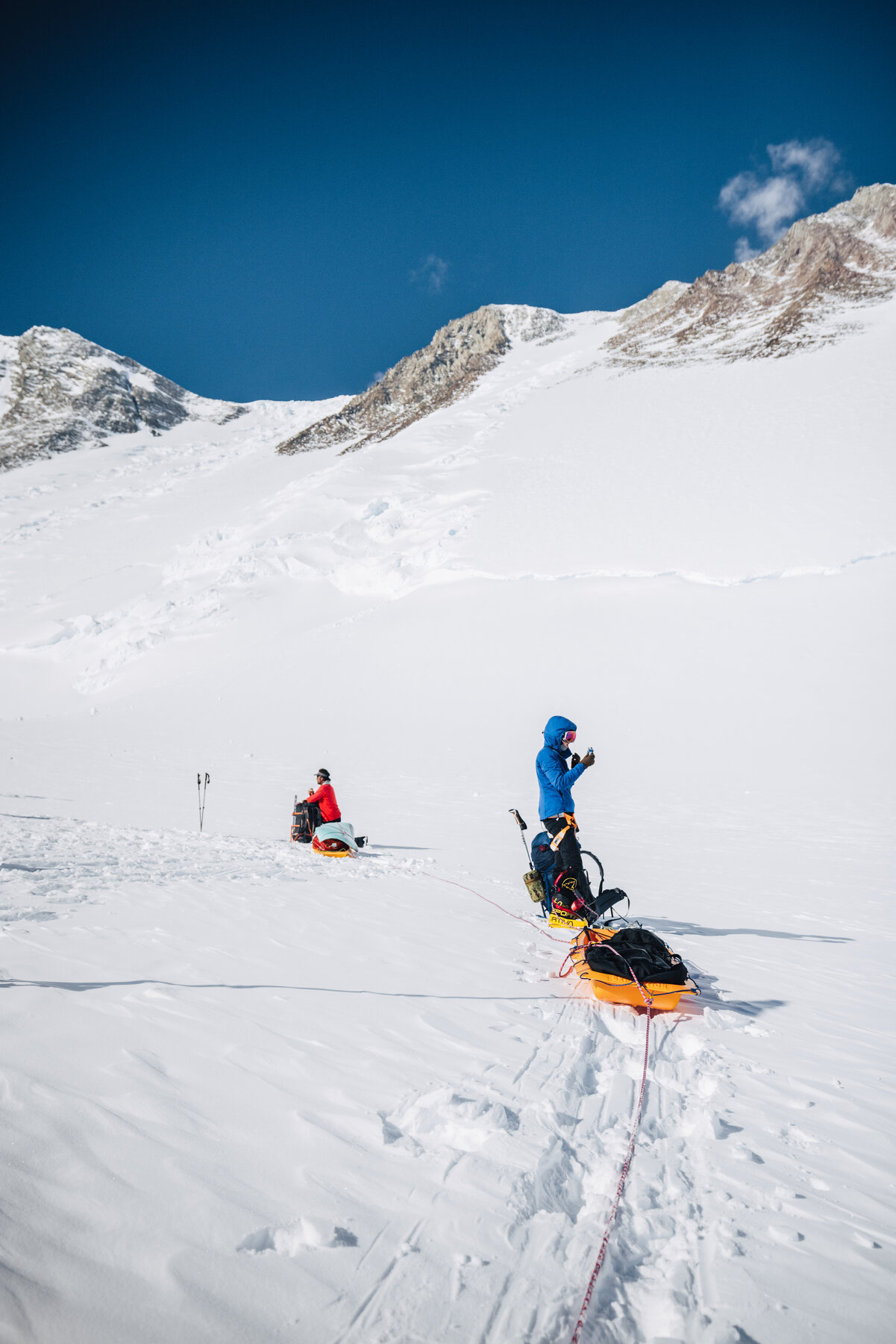 Two climbers hauling orange sleds up the Branscomb Glacier with rocky ridges above