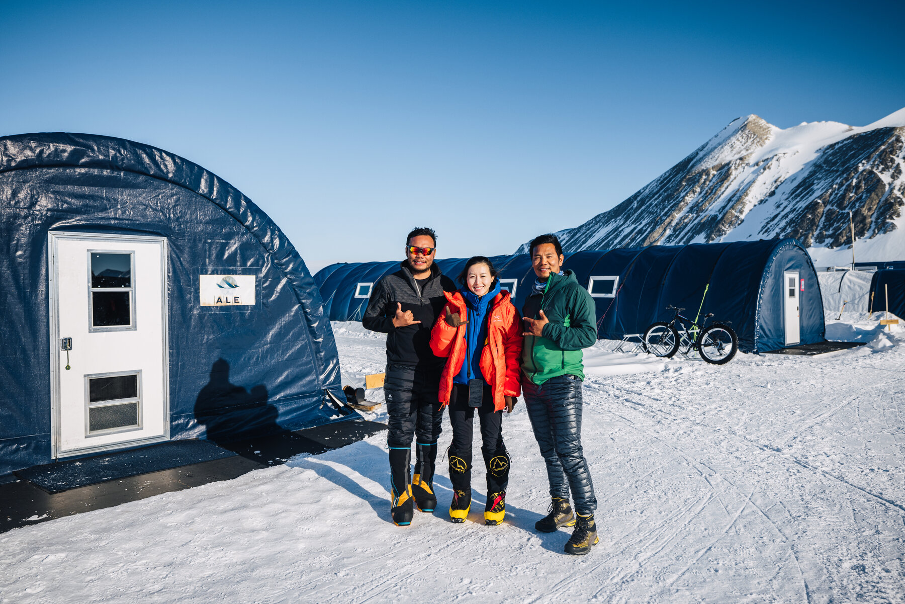 Three climbers posing in front of the blue ALE tents at Union Glacier Camp with fat bikes parked behind and mountains in the distance