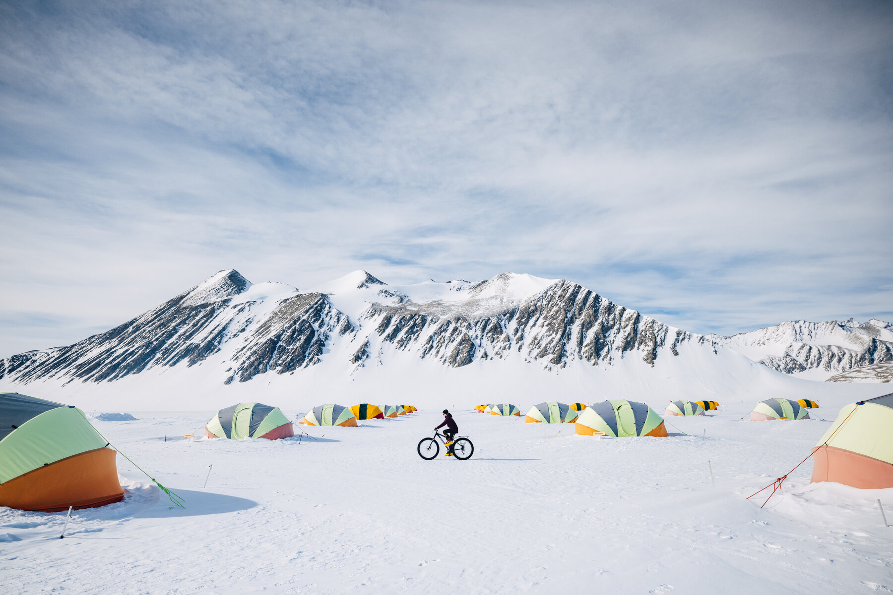 Solo cyclist pedaling a fat tire bike between Union Glacier's colorful tents, snow peaks rising on the horizon