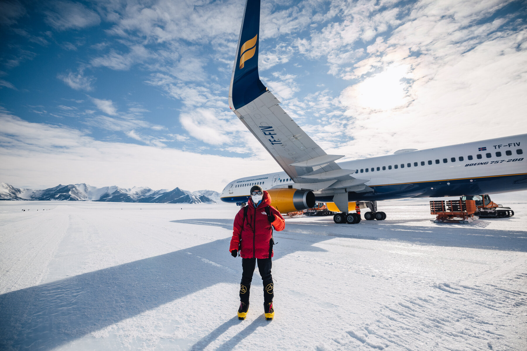 Climber in red jacket standing in front of a large plane on the Union Glacier ice runway with Antarctic mountains behind