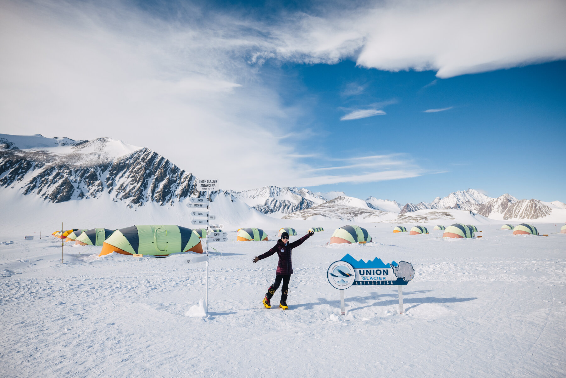 Person with arms outstretched at the Union Glacier Camp welcome sign with tents and mountains behind