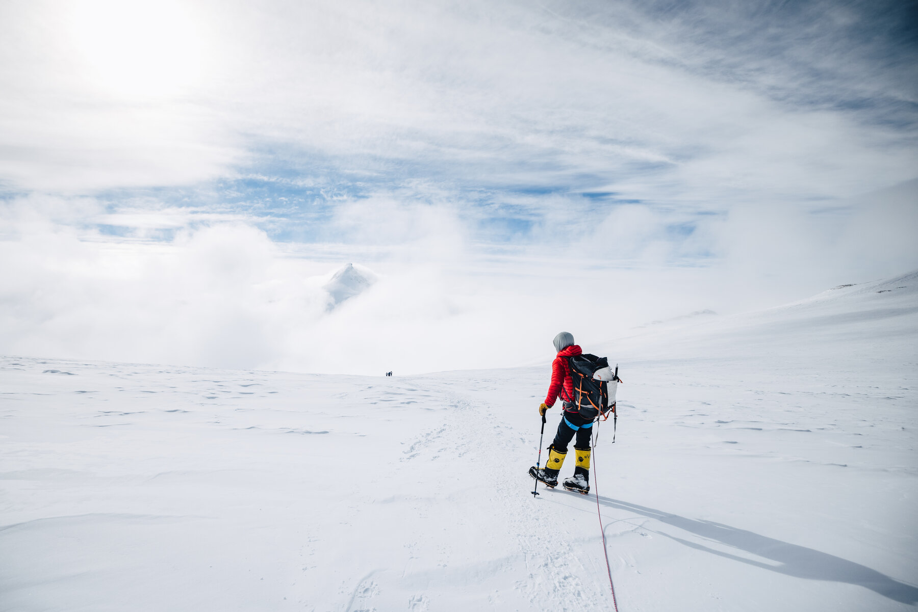 Climber in a red puffy walking through a sea of clouds toward a sub-peak emerging above the cloud layer on summit day
