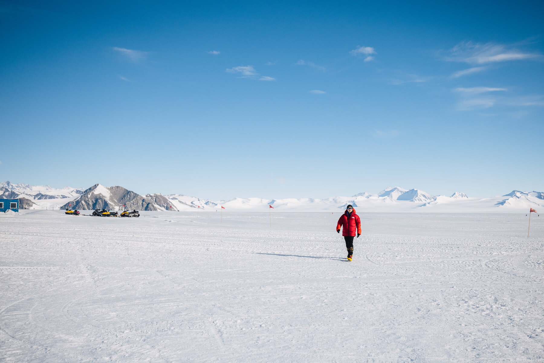 Solo figure walking across the vast white expanse of Union Glacier with camp structures tiny in the distance