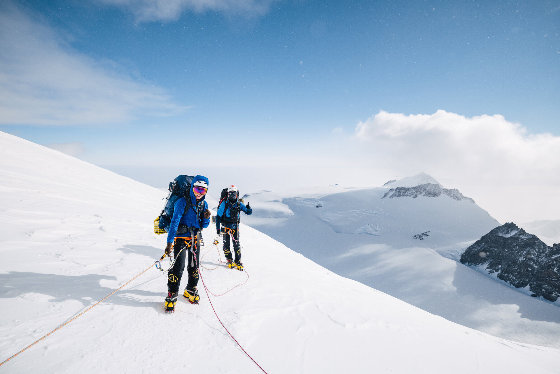 Two roped climbers in helmets and goggles giving a thumbs-up on the upper west face — the Branscomb Glacier and Ellsworth peaks far below